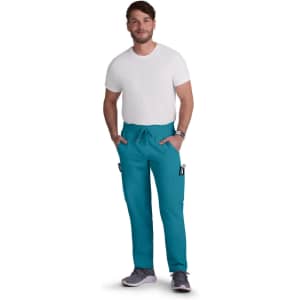 A man wearing teal cargo pants and a white T-shirt, with his hands in the pockets. The pants have a logo.