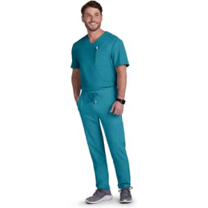 A man in teal scrubs stands smiling, wearing grey shoes and a watch. The outfit includes a top with a pocket and drawstring pants.