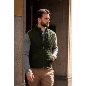 A man wearing a dark green quilted vest over a grey long-sleeve shirt, standing outside a building.