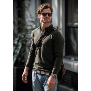A man wearing an olive-green polo shirt with sunglasses, carrying a bag, and dressed in jeans.