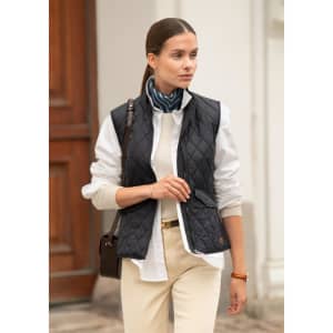 A woman models a navy quilted vest over a white shirt, complemented by a scarf and cream-coloured trousers.