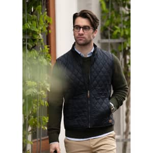 A man in glasses wears a navy quilted vest over a green sweater, walking outdoors with greenery in the background.