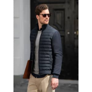 A man wearing a navy quilted jacket and sunglasses, holding a brown leather bag, walking in an urban setting.