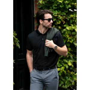 A black polo shirt paired with grey trousers, featuring a logo and styled with a green sweater draped over the shoulder.