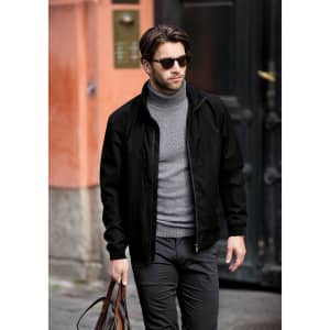 A man in a black jacket and grey turtleneck walks outdoors, carrying a brown bag. The jacket features a logo.