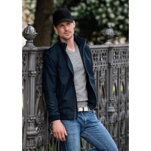 A man in a navy Davenport jacket stands by an ornate fence, wearing a grey shirt and jeans, with a cap.