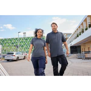 A man and a woman wearing grey work polo shirts, walking together outside a building. The setting features greenery in the background.