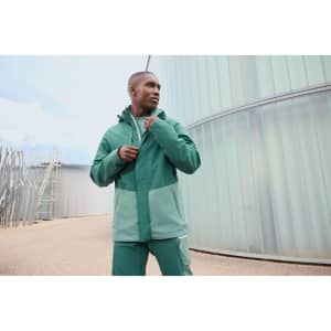 A model wearing a green hardshell rainjacket, featuring contrasting shades and a logo, set in an outdoor industrial environment.