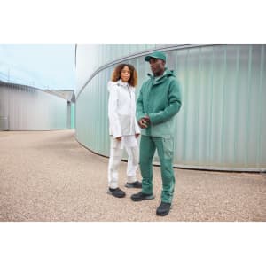 A green hardshell rainjacket is worn by a man, alongside a woman in a white rainjacket, set outdoors.