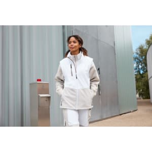 A woman wearing a grey and white softshell vest, walking outdoors near a metal building. The vest has a logo.