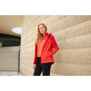 A woman wearing a vibrant red 3-in-1 jacket, standing against a textured stone wall, with a staircase in the background.