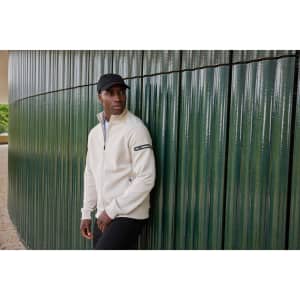 A men's zip jacket in cream, featuring a logo, worn by a model against a textured green wall.