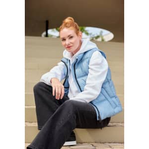 A light blue ladies' winter vest is worn over a white hoodie, paired with black trousers, seated on steps.