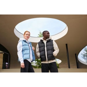 A blue ladies winter vest and a black men's vest are worn by two models outdoors under a circular structure.