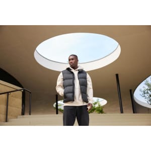 A man wears a black puffer vest over a beige hoodie, posing outdoors under an architectural structure.