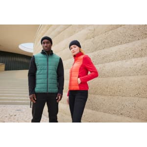 Two individuals stand against a textured wall, wearing a green puffer vest and a red puffer jacket, both in black beanies.