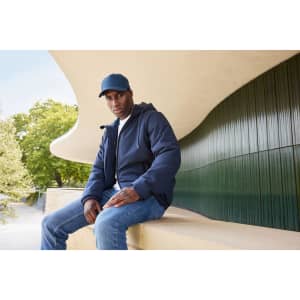 A man sits outside wearing a navy bomber jacket and a matching cap, paired with jeans. The background features greenery and modern architecture.