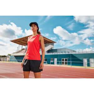 A woman in a red running tank top stands on an athletic track, with a sports facility in the background.