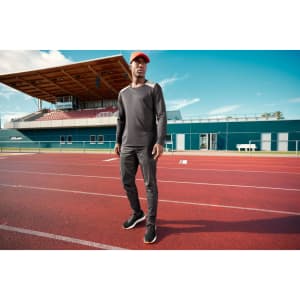 A long-sleeve men's running shirt in grey, paired with black pants and a red cap, on a track field.
