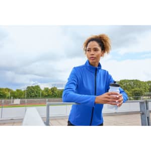 A blue structured fleece jacket is worn by a woman holding a drink bottle, set against an outdoor background.
