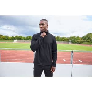 A man wearing a black structured fleece jacket stands outdoors, with a sports track visible in the background.