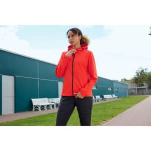 A bright orange softshell jacket with a hood, worn by a woman outdoors, near a green lawn and buildings.