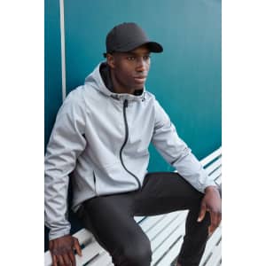 A grey men's sports softshell jacket with a hood, worn by a model seated on a bench, paired with a black cap.