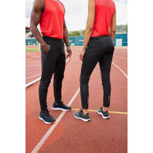 Black track pants worn by two models, paired with red tops and athletic shoes, on an outdoor running track.