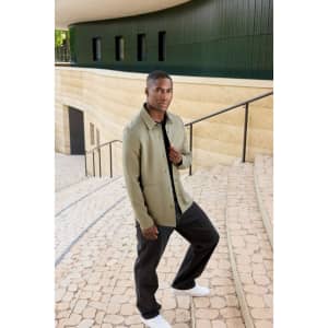 A unisex shirt jacket in olive green, featuring a logo, displayed on a man standing on outdoor steps.