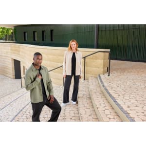 A unisex shirt jacket in olive green and beige worn by models on stone steps, featuring a logo.