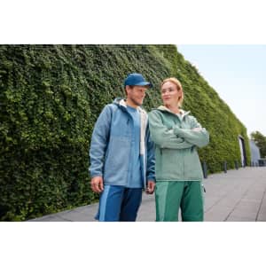 A blue and green knitted fleece jacket worn by a man and a woman, standing outdoors against a green wall.