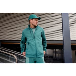 A man wearing a teal workwear fleece jacket and matching cap, standing near an escalator. The jacket has a logo.
