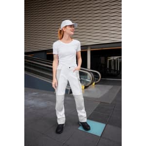 A woman in white workwear pants stands in an outdoor setting, wearing a white t-shirt and cap, with black shoes.