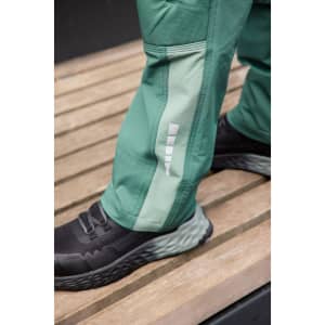 Workwear pants in green with a lighter green panel, featuring reflective details, paired with black footwear on a wooden surface.