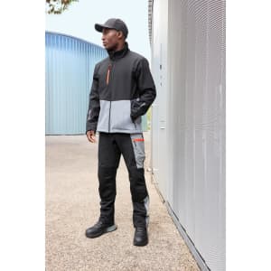 A man wearing a black and grey Workwear Softshell Jacket, paired with black pants and boots, stands against a building.
