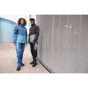 Two models wearing workwear softshell jackets in shades of blue and black, standing against a silver mesh wall.