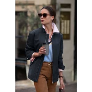 A woman is wearing a dark jacket with a light pink collar, holding a coffee cup in an outdoor setting.