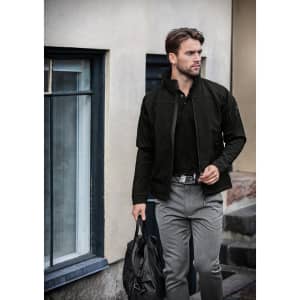 A man wearing a black soft shell jacket, carrying a bag, stands near a building with windows.