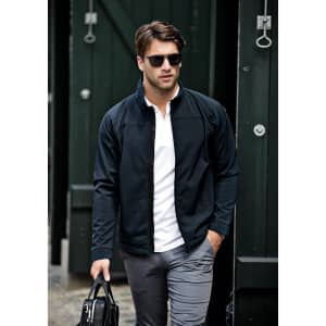 A man in a black soft shell jacket walks confidently, wearing sunglasses and holding a bag.