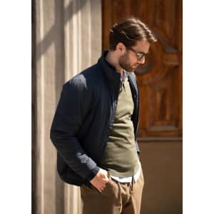 A man wearing a navy jacket over an olive sweater, standing outside next to a wooden door. The jacket has a logo.