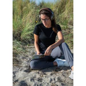 A woman wearing a black t-shirt sits on the sand, listening to music with headphones and reading a book.