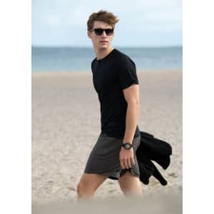 A young man wearing a black T-shirt and grey shorts walks on a beach, with sunglasses and a watch.