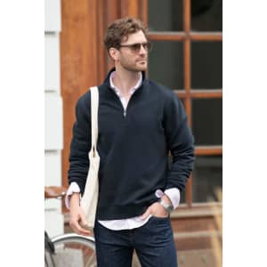 A navy unisex sweatshirt with a zip collar, worn by a man carrying a tote bag. The background features wooden structures.