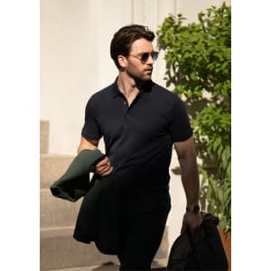 A man wearing a navy polo shirt, holding a green jacket, walking outdoors in sunlight. The image features greenery in the background.