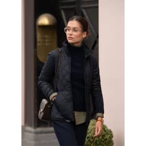 A black quilted women's jacket, styled with a navy top and carrying a handbag, features a sleek design and a logo.