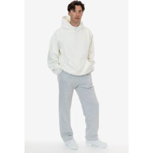 A model wearing a cream hoodie and grey straight leg heavy fleece pants. The pants feature a logo.