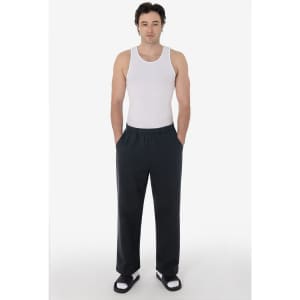 A man wearing a white tank top and black straight leg heavy fleece pants, standing with hands in pockets.