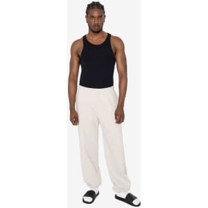 A model wearing cream-coloured heavy fleece sweatpants and a black tank top, paired with sandals. The pants feature a logo.