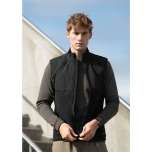 A model wearing a black unisex vest over a long-sleeve brown top, against a stairway and blue sky background.