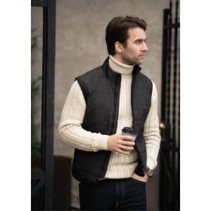 A black men's vest is worn over a cream cable-knit sweater, paired with dark jeans and holding a coffee cup.